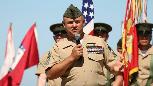 Manuel Diaz, in dress uniform, holds a microphone to address the crowd at his retirement ceremony. The American flag and U.S. Marine Corp flag are behind him, along with other flags and Marines.