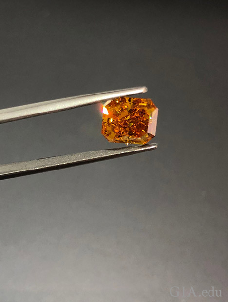 A pair of jewelers tweezers holds an orange diamond.