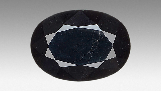 Figure 1. A 22.33 ct oval mixed-cut gadolinite exhibiting a very dark and almost opaque bodycolor and multiple fractures covering the surface. Photo by Shunsuke Nagai.