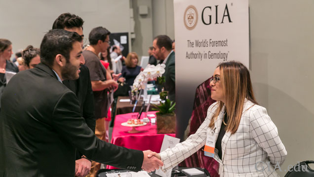 A man and woman shake hands during a career fair event. In the background is a GIA poster.