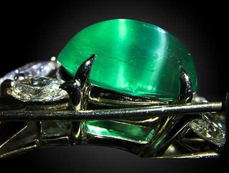 Figure 2. Side view of the emerald ring. Parallel tube inclusions are oblique to the pavilion. The white reflection on the right is caused by the inclusions near the surface of the curved cabochon, while the greenish reflection on the left is caused by the reflection off of the bottom face of the cabochon. Photo by Shunsuke Nagai. Figure 2. Side view of the emerald ring. Parallel tube inclusions are oblique to the pavilion. The white reflection on the right is caused by the inclusions near the surface of the curved cabochon, while the greenish reflection on the left is caused by the reflection off of the bottom face of the cabochon. Photo by Shunsuke Nagai.
