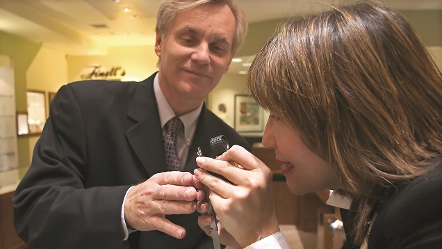A male retail jeweler helps a woman look at a diamond through a loupe. A male retail jeweler helps a woman look at a diamond through a loupe.