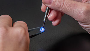 Close up of hands holding a loose stone with tweezers in one hand, and using the iD100 probe to highlight the stone in the other on a black background. 