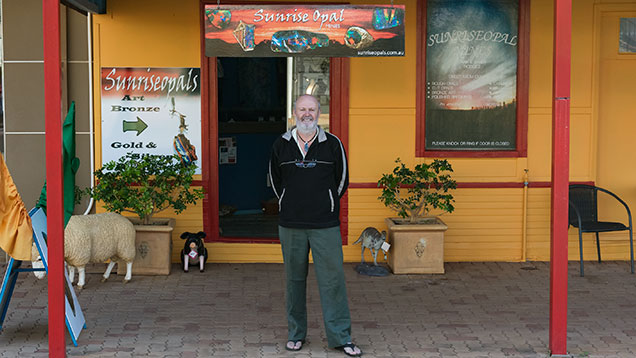 Man outside store. Mark Hodges（马克·霍奇斯）自豪地站在他的 Quilpie（奎尔派）店 — Sunrise Opals（日出蛋白石）外。 对于 Mark（马克）和他的妻子 Sandy（桑迪）来说，蛋白石切割和创造珠宝的艺术满足感使得他们开始涉足零售业，出售自己的作品。 摄影：Andrew Lucas（安德鲁·卢卡斯）/GIA。. 店舗の外に立つ男性