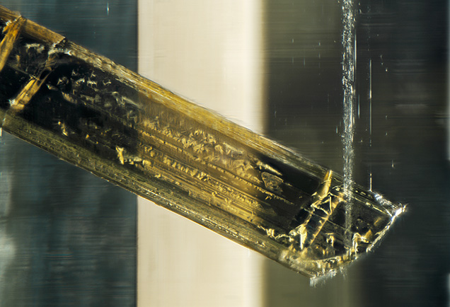 Morphological features of the tourmaline crystal included in the heliodor are visible; striations and triangular growth marks are present. A band of fluid inclusions is visible near the tip of the schorl tourmaline. Photomicrograph by Rhiana Elizabeth Henry; field of view 2.90 mm. Gift of Mark Mauthner, GIA Museum no. 37772.