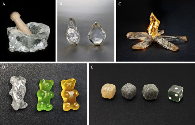 Figure 7. Rough diamonds resembling objects: mortar, 1.95 ct, with a pestle carved from a toothpick (A); raindrops, 3.23 ct each (B); campfire flame, 2.70 ct, backlit using orange light and placed with raw baguettes from figure 3 as firewood (C); gummy bear, 9.18 ct (D; colored gummy bears for comparison created using Adobe Photoshop Generative AI); fibrous diamond cuboids resembling dice, 22.45, 22.47, and 23.64 ct, respectively, shown with a superimposed 10 mm die (E). Photos by Evan M. Smith.