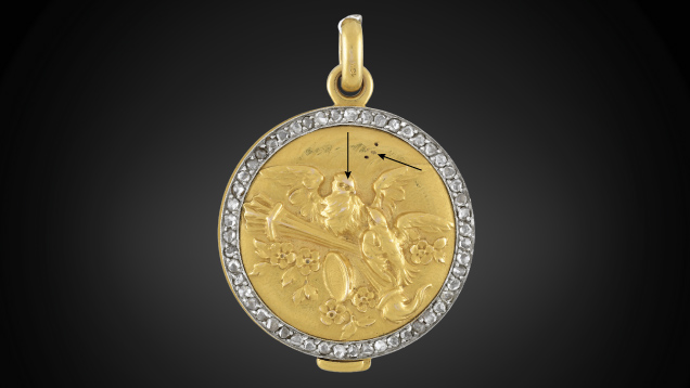 Figure 2. In this gold-plated pendant submitted as 18K solid gold, the base layer tested 10K and the top layer tested 18K. Arrows show where the gold plating has rubbed off due to use. Photo by Diego Sanchez. Figure 2. In this gold-plated pendant submitted as 18K solid gold, the base layer tested 10K and the top layer tested 18K. Arrows show where the gold plating has rubbed off due to use. Photo by Diego Sanchez.