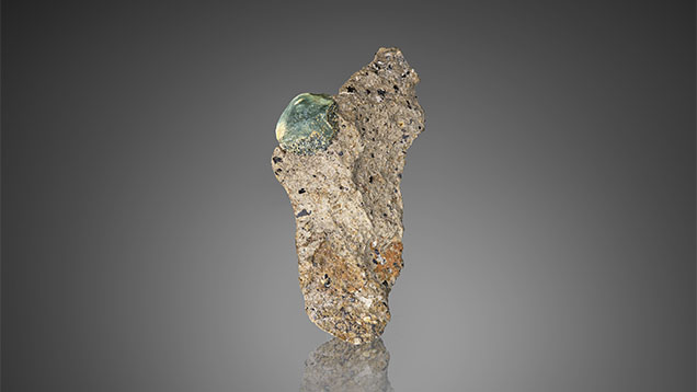 Figure 1. A gem-quality <em>in situ</em> French Bar sill primary sapphire (9 mm long) partially embedded in basaltic trachyandesite rock (sample RK-FBS 001). Photo by Jeff Mason.