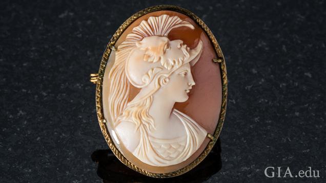 A side profile of Athena shows the top of her toga-like dress and a fanned helmet with a portrait of her father Zeus on it. The carving is ivory with shades of orange and background of the shell is taupe.