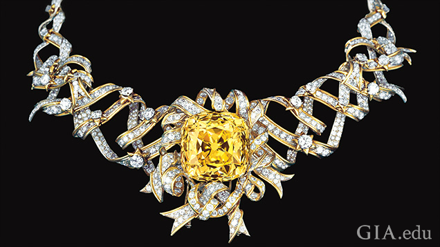 大きなロゼット カット イエロー ダイヤモンドが、ダイヤモンドでできたリボンに囲まれて置かれている。