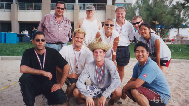 A group of 11 casually dressed students pose for a group photo in the sand of the volleyball court at GIA’s Carlsbad campus.