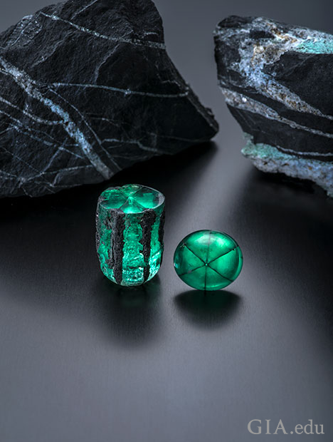 A barrel shaped piece of traphiche emerald rough and a cabochon of it sit in front of two large chucks of shale rock.