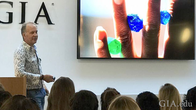 Brian Cook stands at a podium to the left of a big screen with an image of an upheld hand with brightly colored green and blue gems held between four fingers.