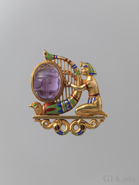 A kneeling figure clad in Egyptian headdress, arm bands, and loincloth playing a falcon-headed harp set with an oval amethyst carved as a scarab. The figure and harp rest on a plinth supported by two coiled snakes. Green, blue, and red enamel accent the h