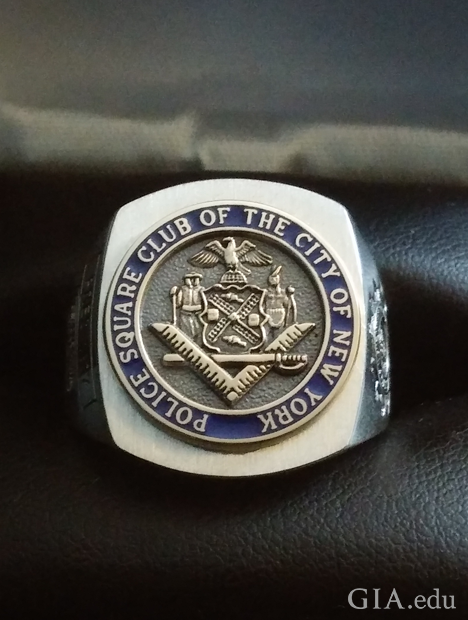 A square shaped ring with a circular center with raised symbols. It is engraved with “Police Square Club of the City of New York.”