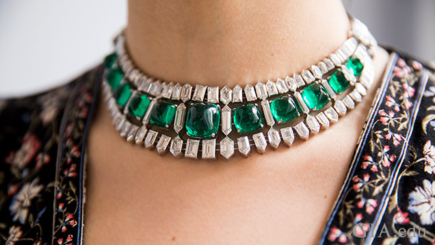 A wide choker-style necklace of a row of emeralds with rows of diamonds above and below the emeralds.