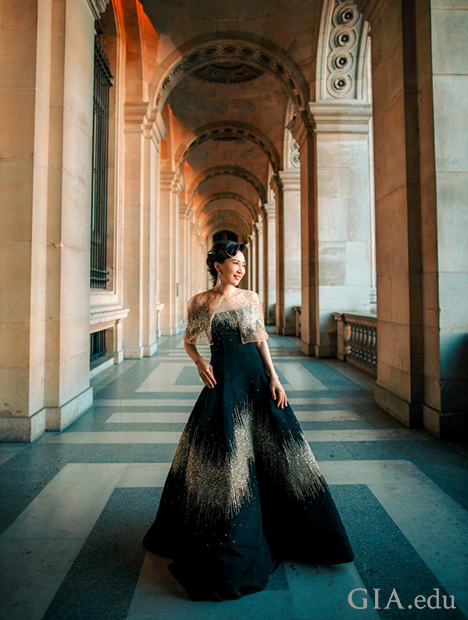 Woman stands in a long, ornate hallway, dressed in formal gown.