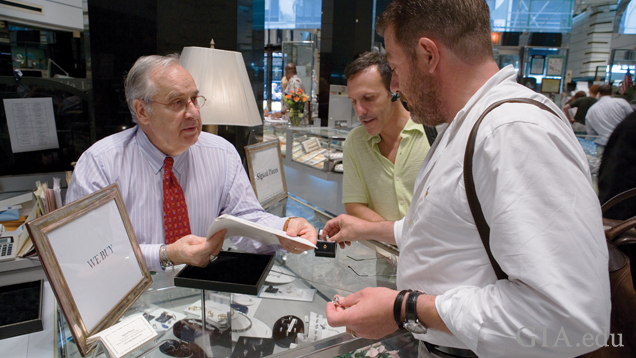 Three men look at diamond jewelry and paperwork at a jeweler’s counter.