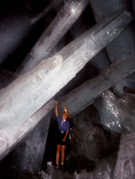 Huge crystals tower above and around woman in a dark cave. 
