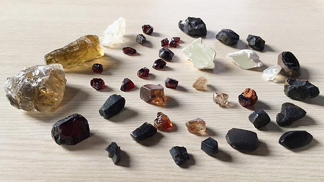 Figure 1. Gem materials from southeast Vietnam. The three largest yellow and white samples on the far left are feldspar, while the brown, orange, and near-colorless samples in the center are zircon and the two greenish white samples near the top right are hyalite opal. The rest of the dark stones are sapphires, garnets, augites, and brown peridot. Photo by Le Ngoc Nang; courtesy of Tran Ngoc Vien.