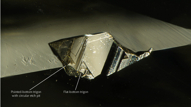Figure 2. Flat-bottom and pointed-bottom trigons (surface features restricted to an octahedral crystal face) preserved on an unpolished portion of the diamond. A circular etch pit overprinting a pointed-bottom trigon indicates a late-stage diamond dissolution event that postdated the formation of the trigon. Photomicrograph by Matthew Hardman; field of view 1.26 mm.