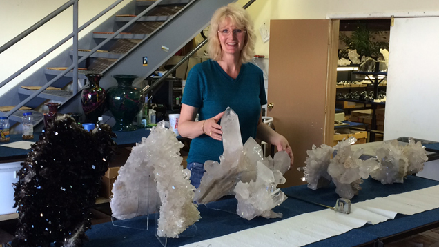 Terri Ottaway stands behind a table with several very large quartz crystal specimens are lined up on it. Some of them come up to her waist height.
