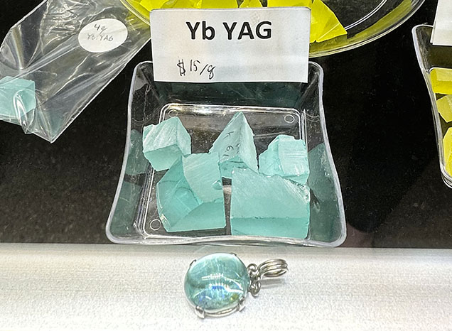 Figure 3. Greenish blue rough and cut ytterbium-doped YAG. The cabochon in front is 14 × 10 mm. Photo by Lisa Kennedy; courtesy of Turtle’s Hoard.