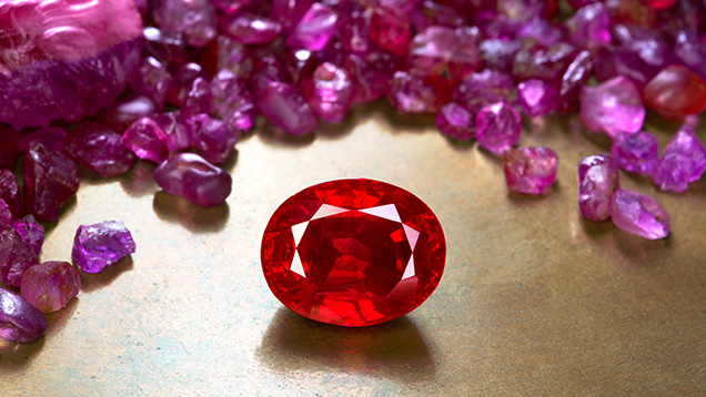 Figure 1. A faceted heated ruby from Mogok, Myanmar, weighing more than 3 ct, stands out against a backdrop of untreated ruby rough from the same origin. The striking appearance of Mogok ruby is highly sought after. Photo by Wimon Manorotkul; faceted ruby courtesy of Kiarttichatra Intarungsee.