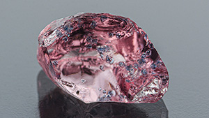 Figure 1. Left: This 3.07 ct purplish pink “dragon” garnet, as seen in daylight-equivalent lighting, displays eye-visible blue apatite inclusions. Right: Red fluorescence in the garnet due to chromium, as seen using long-wave UV. Photos by Jessa Rizzo; courtesy of Ravenstein Gem Co.