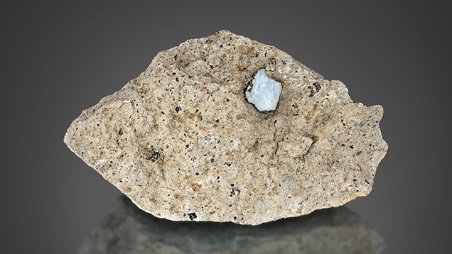 Figure 11. A sapphire (8 mm in length) in French Bar sill matrix rock showing a thin biotite (phlogopite) coating (RK-FBS 003). To be preserved as an attractive mineral specimen, enough matrix surrounding the sapphire is removed. Photo by Jeff Mason.